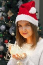 Woman in santa hat with christmas lights in her hands looks at camera in room with xmas tree, festive holiday shine Royalty Free Stock Photo