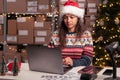 Woman in Santa hat preparing fast xmas delivery uses laptop computer. Workshop warehouse on Christmas and delivering