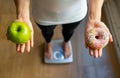 Woman on scale measuring weight holding apple and donuts choosing between healthy or unhealthy food Royalty Free Stock Photo