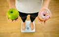 Woman on scale measuring weight holding apple and donuts choosing between healthy or unhealthy food Royalty Free Stock Photo