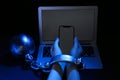 Woman shackled with ball and chain holding smartphone near laptop on dark background, closeup. Internet addiction