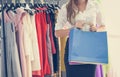 Woman shopping and holding shopping bag in women fashion clothing store with colorful women`s dresses on hangers in a retail shop