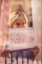 Woman staying in flat in front of window holding a sheet of paper with title self-isolation. People and healthcare concept