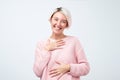 Woman with short dyed hair wearing pink sweater smiling broadly and keeping hand on her heart