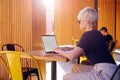 Woman with short hair working on laptop, screen space for design layout. Sitting outside in modern cafe yellow wall, sunny day