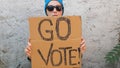 Woman Shows Cardboard With Go Vote Sign On Brick Wall Urban Background. Voting Concept. Make Political Choice, Elections