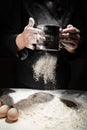 woman sifts flour through sieve onto table with eggs on black background.