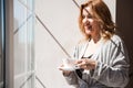 A woman in a silver dressing gown stands near the window with a cup of drink in her hands. The weather is good outside, the sun is