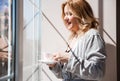 A woman in a silver dressing gown stands near the window with a cup of drink in her hands. The weather is good outside, the sun is