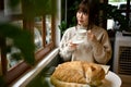 A woman sipping tea or coffee at a table by the window while her cat is sleeping on a table Royalty Free Stock Photo