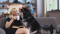 Woman sits on the couch, reads a book, drinks coffee and talks to the husky while her husband is cooking something in