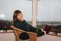 Woman sits in a cozy wicker chair on a light covered veranda in the mountains, enjoys relaxing, smiling, stretching out her legs