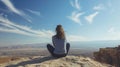 A woman sits crosslegged on a rock back to the camera as gazes towards the endless blue sky and endless stretch of the