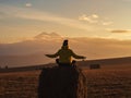 A woman sits on a haystack with her arms spread apart against the backdrop of Elbrus in the clouds at sunset. View from Royalty Free Stock Photo