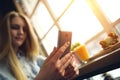 Woman sits near a large window in a modern cafe at a table and writes something in the smartphone, the focus is positioned on a sm