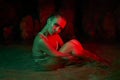 Woman sits in water at night, illuminated by green red light. Female model in aquatic environment, creative lighting