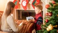 Young woman sitting in armchair in living room and giving box with Christmas present to her little son Royalty Free Stock Photo