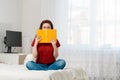 A woman is sitting on the bed and covers her face with a yellow book. A bright room and a computer in the background. Concept of