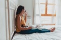 Woman sitting on the bed looking out the window drinking a cup of coffee with her dog Jack Russell terrier lying in her legs