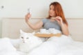 A woman is sitting in bed with a mobile phone in her hands. A white cat lies at the feet of the mistress with a cold Royalty Free Stock Photo