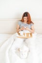 A woman is sitting in bed with a mobile phone in her hands. A white cat lies at the feet of the mistress with a cold. A Royalty Free Stock Photo