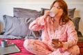 A woman is sitting on a bed with a phone during a covid-19 flu virus illness. Girl in pink pajamas drinks from a cup