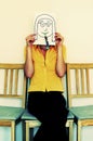 A woman sitting on a chair covering her face with a drawing of a girl with spectacles and blowout. Conceptual image shot
