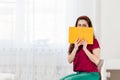 A woman is sitting on the chair and covers her face with a yellow book. A bright room in the background. Concept of education and