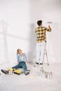 Woman sitting on floor while boyfriend standing on ladder with pink paint roller