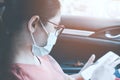 Woman sitting in the front of a car at shotgun seat by wears medical face mask and holding smarts phone in her hand. Safety and