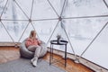 A woman is sitting in a geo dome glamping tent in a bag chair and looking out at the winter snow-covered nature and drinks coffee Royalty Free Stock Photo