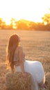 Woman sitting on hay stack walking in summer evening, beautiful romantic girl with long hair outdoors in field at sunset Royalty Free Stock Photo