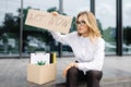 Woman sitting outdoor with poster Act Now next to box of stuff . Beautiful female protester at strike against unemployment Royalty Free Stock Photo