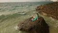 Woman sitting on rock of sea reef stone, stormy cloudy ocean. Blue swimsuit dress tunic. Concept resort coastline Royalty Free Stock Photo