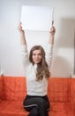 Woman sitting on a sofa and holding outstretched empty sheet of paper with copy spac