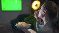 Woman sitting on a sofa in the living room in the evening and watching a green TV screen mockup. The woman is infatuated