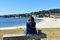 Woman sitting on a stone bench in a beach promenade. Long hair, blue clothes. Bright sand, turquoise water. Sunny. Galicia, Spain.