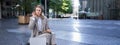 Woman sitting on a street with laptop and headphones plugged in, taking notes. Corporate worker attend online team