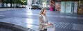 Woman sitting on a street with laptop and headphones plugged in, taking notes. Corporate worker attend online team