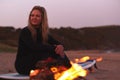 Woman Sitting On Surfboard By Camp Fire On Beach As Sun Sets Behind Her Royalty Free Stock Photo