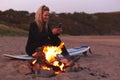 Woman Sitting On Surfboard By Camp Fire On Beach Using Mobile Phone As Sun Sets Behind Her Royalty Free Stock Photo