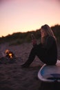 Woman Sitting On Surfboard By Camp Fire On Beach Using Mobile Phone As Sun Sets Behind Her Royalty Free Stock Photo