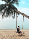 Woman sitting on swing hanging on palm tree and looking at calm blue ocean on paradise sandy beach