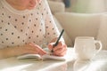 Woman sitting at the table, writing in the notebook and drinking coffee in nice light home interior. Working at home. Freelancer.