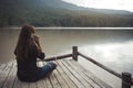 A woman sitting and talking on mobile phone on an old wooden pier by the river with sky and mountain background Royalty Free Stock Photo