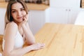 Woman is sitting at wooden table in front of white cozy kitchen, is smiling and looking at camera Royalty Free Stock Photo