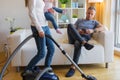 Woman with small child doing housekeeping while man sitting in couch