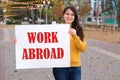 A woman smiles and holds a poster with the text Work Abroad