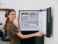 Woman smiling with teeth looking into camera in kitchen at home opened freezer empty with ice inside, home refrigerator