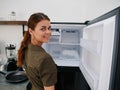 Woman smiling with teeth looking into camera in kitchen at home opened freezer empty with ice inside, home refrigerator